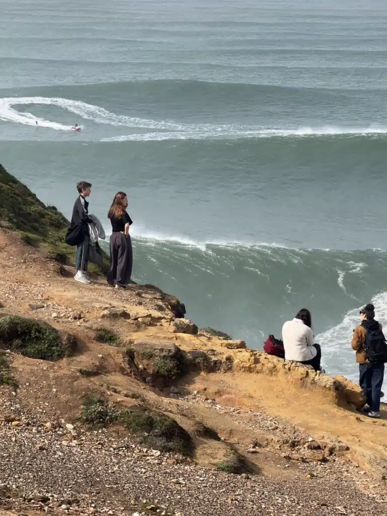 Jet ski towing across a big swell at Nazaré while people watch from the cliff